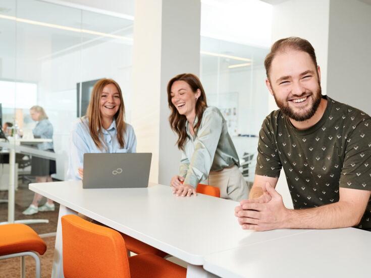 Drei Kolleg:innen lachen und arbeiten gemeinsam an einem hellen Büroarbeitsplatz