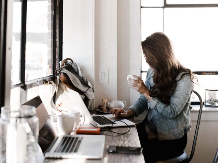 Eine Frau mit langen Haaren sitzt im Büro an einem Laptop und hält eine Tasse Kaffee in der Hand