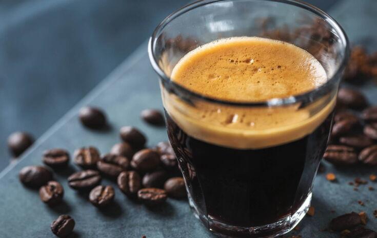 Ein transparentes Schnapsglas mit einem Corretto darin vor einem blauen Hintergrund mit Kaffeebohnen