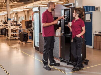 Zwei Mitarbeiter trinken Kaffee neben Kaffeevollautomat in einer Werkstatt.