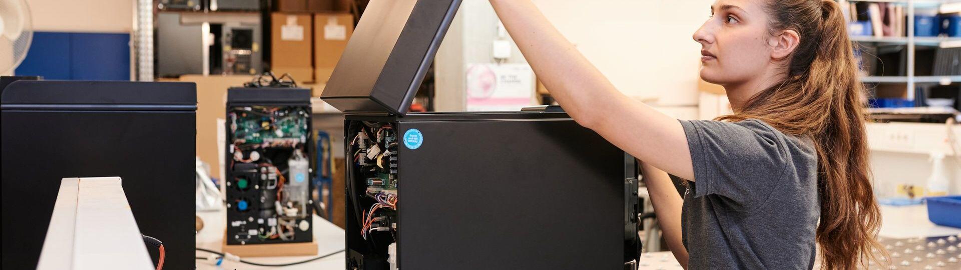 Fachkundige Technikerin klappt einen hochwertigen Kaffeevollautomaten in einer modernen Montagehalle auf.