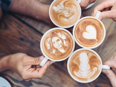 Vier Kaffeetassen mit Latte Art und einem Porträt im Schaum