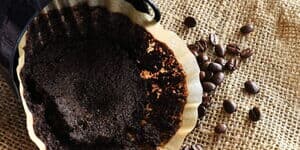 Gebrauchter Kaffeefilter mit feuchtem Kaffeesatz in einem Filterhalter, daneben verstreute Kaffeebohnen auf grobem Jutestoff