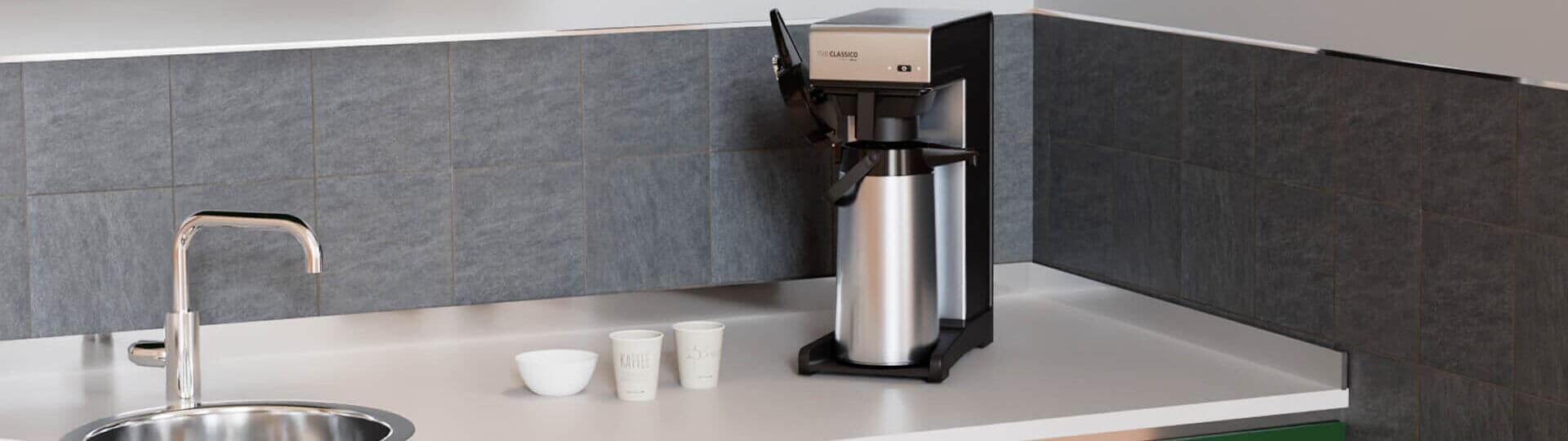 Filterkaffeemaschine auf einer Küchenarbeitsplatte, umgeben von grünen Schränken und schwarzer Fliesenrückwand.