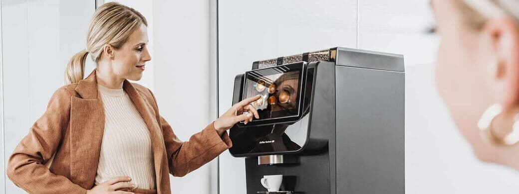 A woman in elegant office clothing operates a fully automatic coffee machine from Kaffee Partner in the office. Another woman can be seen slightly out of focus in the foreground.