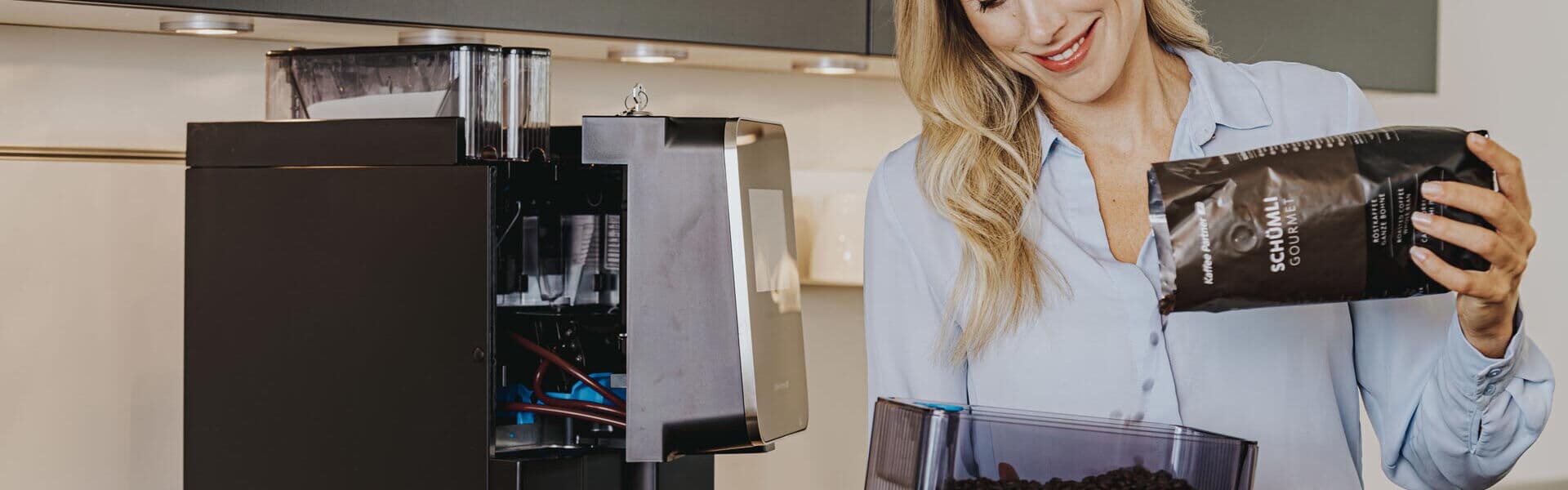 Frau steht lächelnd vor einer Küchenzeile, auf dem sich ein offener Kaffeevollautomat steht, und füllt den Produktbehälter mit Kaffeebohnen auf, den sie in der Hand hat