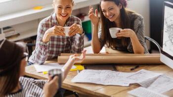 Drei junge Frauen sitzen an einem Schreibtisch und trinken Kaffee