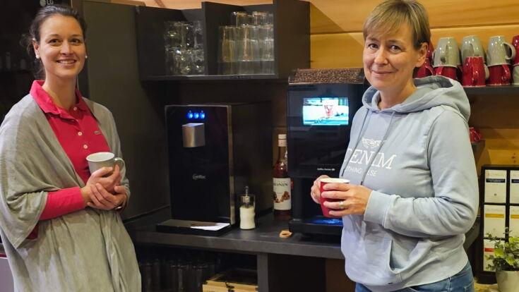 Zwei Kolleginnen genießen eine Kaffeepause neben einem Kaffeevollautomaten und Wasserspender im Büro von AWEWA-Coaching
