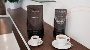Zwei Kaffeepackungen von Kaffee Partner – Espresso Superiore und Schümli Gourmet – stehen nebeneinander auf einem dunklen Holztresen. Davor jeweils eine weiße Tasse mit frisch gebrühtem Kaffee auf Untertasse und Löffel.