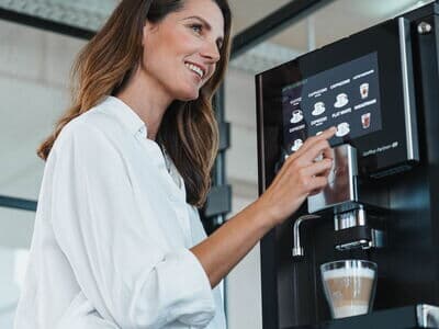Frau zieht sich einen Latte macchiato aus einem modernen Kaffeevollautomaten während sie zur Seite guckt