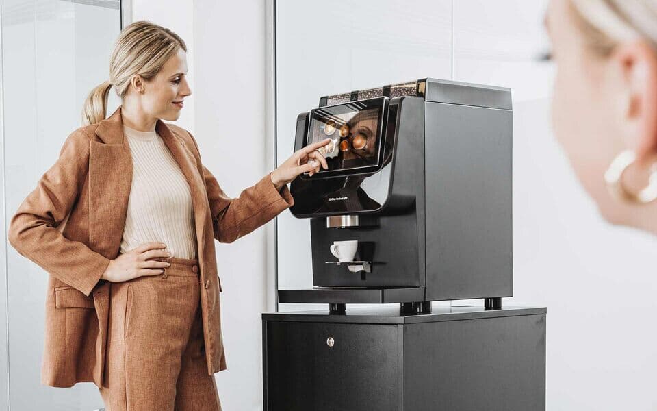 Eine Frau in eleganter Bürokleidung bedient einen Kaffeevollautomaten von Kaffee Partner im Büro. Eine weitere Frau ist im Vordergrund leicht unscharf zu sehen.