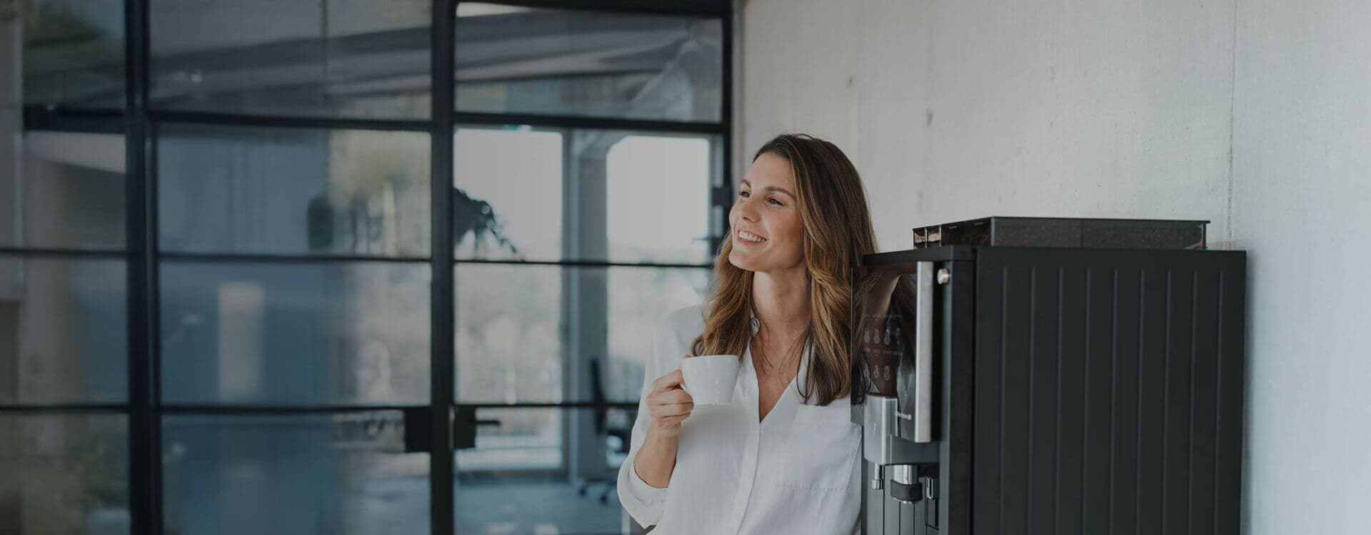 Eine lächelnde Frau lehnt entspannt an einem schwarzen Kaffeevollautomaten im Büro und hält eine weiße Tasse in der Hand. Im Hintergrund sind Glaswände und ein Besprechungsraum sichtbar.