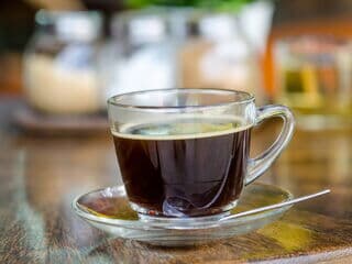 Glastasse mit schwarzem Kaffee steht auf einem Holztisch in gemütlicher Café-Atmosphäre.