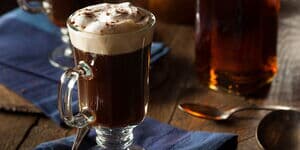 Ein Glas Irish Coffee mit cremiger Sahnehaube, serviert auf einem blauen Stofftuch, daneben ein Löffel auf einem rustikalen Holztisch