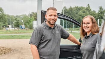 Zwei Servicemitarbeitende stehen vor einem Firmenfahrzeug mit offener Tür und lächeln in die Kamera. Sie tragen Arbeitskleidung mit Firmenlogo.