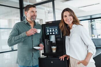 Eine lächelnde Frau und ein Mann mit einer Kaffeetasse in der Hand stehen zwischen einem Kaffeevollautomaten.