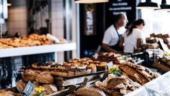 Auslage ein Bäckerei mit Brot und Kuchen. Im Hintergrund stehen zwei Mitarbeiter hinter dem Tresen.