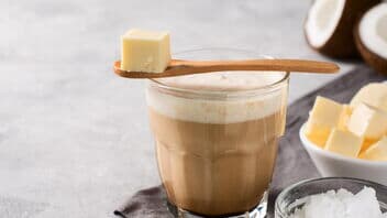 Ein Glas Bulletproof Coffee mit cremiger Schaumkrone, davor Butterwürfel, MCT-Öl und eine Kokosnusshälften.