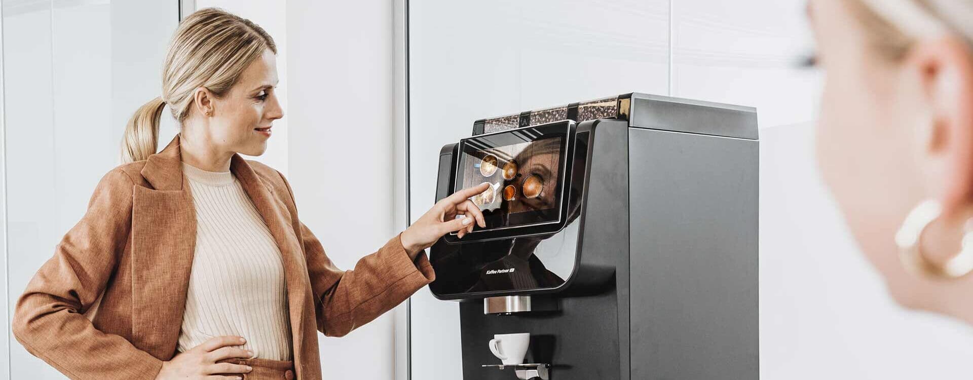 Eine Frau in eleganter Bürokleidung bedient einen Kaffeevollautomaten von Kaffee Partner im Büro. Eine weitere Frau ist im Vordergrund leicht unscharf zu sehen.