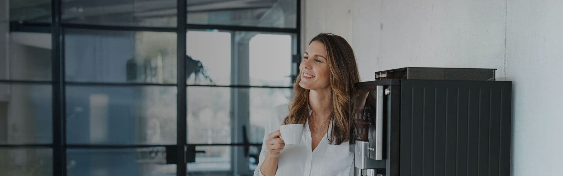 Eine lächelnde Frau lehnt entspannt an einem schwarzen Kaffeevollautomaten im Büro und hält eine weiße Tasse in der Hand. Im Hintergrund sind Glaswände und ein Besprechungsraum sichtbar.