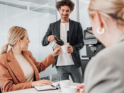 Ein Mann gießt Kaffee aus einer Kanne in eine Tasse einer Kollegin, während sie an einem Tisch in einem modernen Büro sitzt. Eine weitere Kollegin sitzt im Vordergrund, während im Hintergrund ein Kaffeevollautomat zu sehen ist.