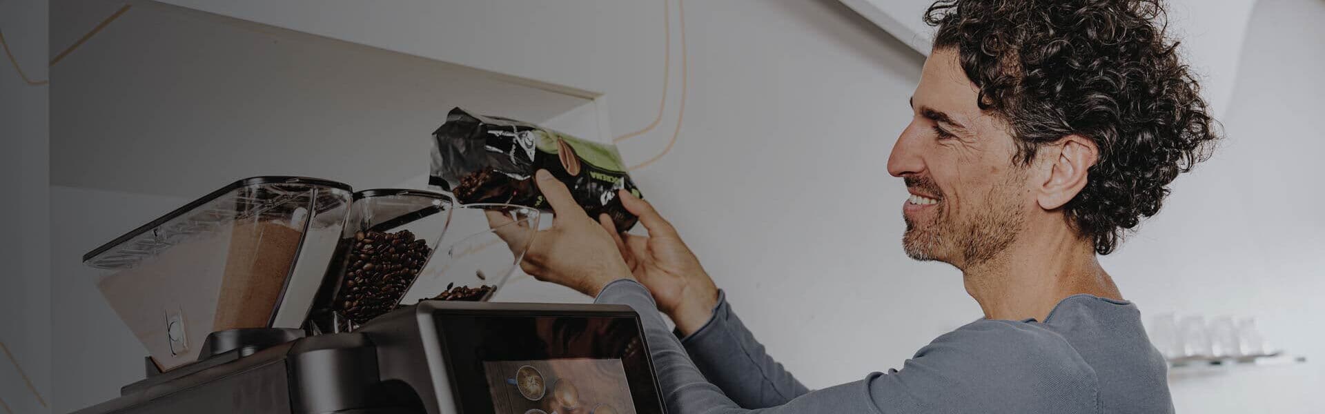 Ein lächelnder Mann füllt Kaffeebohnen in den Behälter eines modernen Kaffeevollautomaten in einem Büro.