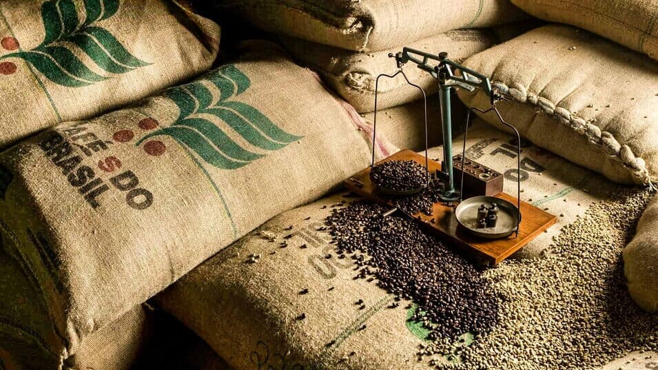 Kaffeebohnen auf Waage umgeben von weiterem Kaffeebohnen platziert auf Jutesäcken mit brasilianischem Aufdruck