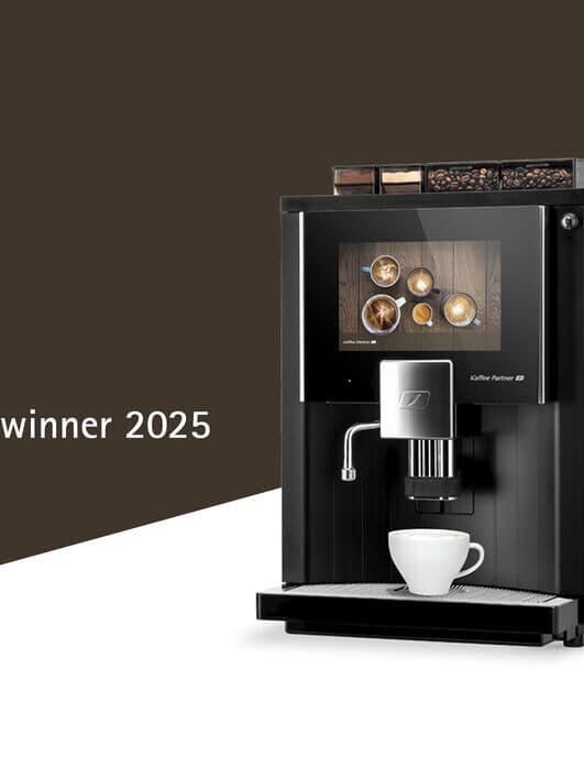 Kaffeevollautomat von Kaffee Partner mit moderner Touchscreen-Oberfläche, ausgezeichnet mit dem Red Dot Design Award 2025. Auf dem Bildschirm sind verschiedene Kaffeespezialitäten zu sehen. Links daneben das Red Dot Winner Logo auf braunem Hintergrund.