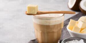Ein Glas Bulletproof Coffee mit cremiger Schaumkrone, davor Butterwürfel, MCT-Öl und eine Kokosnusshälften.