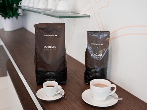 Zwei Kaffeepackungen von Kaffee Partner – Espresso Superiore und Schümli Gourmet – stehen nebeneinander auf einem dunklen Holztresen. Davor jeweils eine weiße Tasse mit frisch gebrühtem Kaffee auf Untertasse und Löffel.