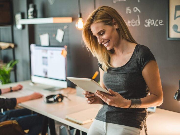 A smiling woman stands in the office with a tablet in her hand