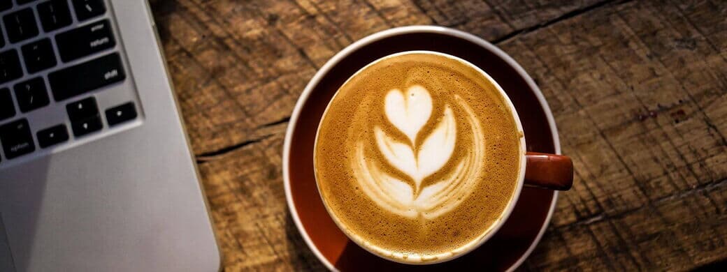 Eine Tasse Kaffee mit einer Blume aus Milchschaum steht neben einem Laptop auf einem Holztisch