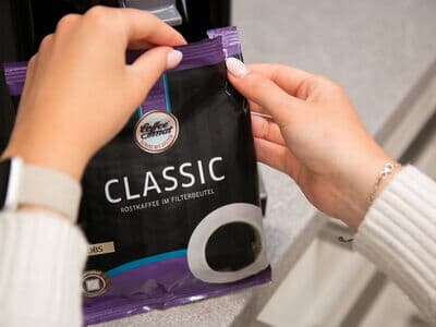 Person hält eine Packung 'Coffee Mat Classic' Röstkaffee in Filterbeuteln vor einer Kaffeemaschine in der Küche.