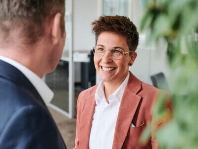 Eine Sales Managerin mit Kurzhaarschnitt, Brille und Blazer führt ein Gespräch mit einem Geschäftspartner im Büro. Sie lächelt freundlich.