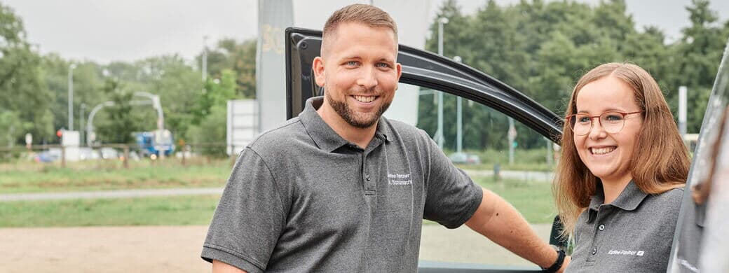 Zwei Servicemitarbeitende stehen vor einem Firmenfahrzeug mit offener Tür und lächeln in die Kamera. Sie tragen Arbeitskleidung mit Firmenlogo.