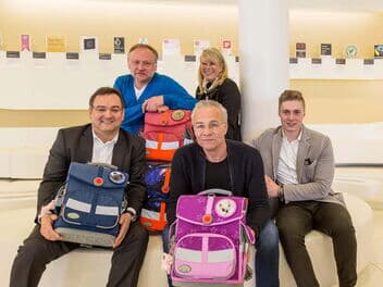 Mehrere Personen sitzen zusammen und halten Schulrucksäcke.