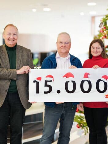 Fünf Personen stehen vor einem weihnachtlich geschmückten Hintergrund mit einem Weihnachtsbaum. Sie halten ein Schild mit der Aufschrift „15.000“, auf dem kleine Weihnachtsmützen dekorativ angebracht sind.