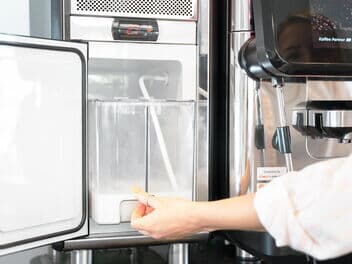 Nahaufnahme eines Kaffeevollautomaten von Kaffee Partner mit angeschlossenem Milchschlauch und Milchkühler, Hand zeigt auf den Milchkühler