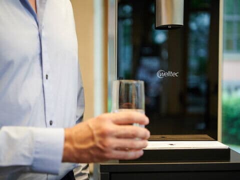 Die Glasfläche des Wasserspenders mit welltec Logo ist hinter einer Hand mit einem Glas Wasser zu sehen