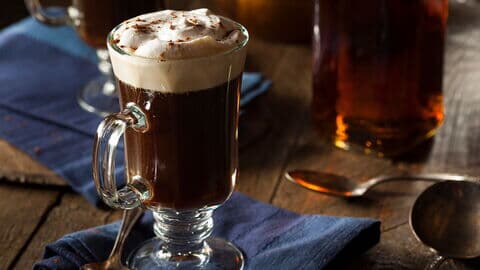 Ein Glas Irish Coffee mit cremiger Sahnehaube, serviert auf einem blauen Stofftuch, daneben ein Löffel auf einem rustikalen Holztisch