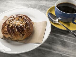 Ein schwedisches Gebäckstück liegt auf einer beigen Serviette auf einem weißen Teller und daneben steht ein schwarzer Kaffee in einer blauen Tasse mit gelbem Unterteller und einem silbernen Löffel.