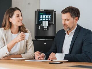 Ein Frau und ein Mann sitzen an einem Tisch. Die Frau guckt den Mann an der etwas aufschreibt. Im Hintergrund ist ein Kaffeevollautomat zu sehen.