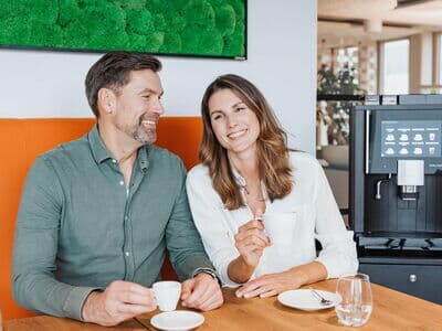 Mann und Frau trinken Kaffee neben einem Kaffeevollautomat während sie nebeneinander an einem Tisch sitzen