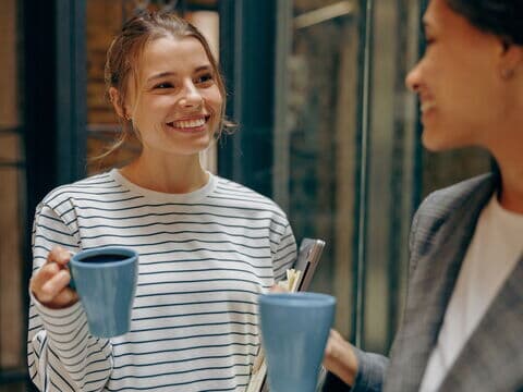 Zwei Frauen unterhalten sich lächelnd bei einer Kaffeepause im Büro.