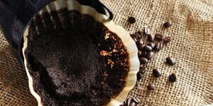 Gebrauchter Kaffeefilter mit feuchtem Kaffeesatz in einem Filterhalter, daneben verstreute Kaffeebohnen auf grobem Jutestoff