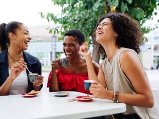 Drei lachende hispanische Frauen sitzen im Freien an einem Tisch und trinken Kaffee