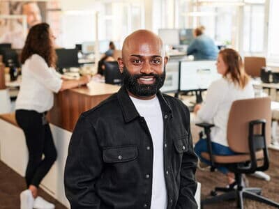Ein freundlicher Mitarbeiter mit Bart steht im Büro und lächelt in die Kamera. Im Hintergrund arbeiten seine Kolleginnen und Kollegen an Computern.