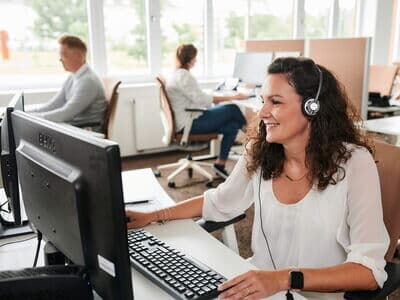 Eine freundliche Mitarbeiterin mit Headset telefoniert an ihrem Arbeitsplatz im Büro. Sie sitzt vor einem Computer und arbeitet konzentriert im Kundenservice.