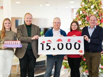 Fünf Personen stehen vor einem weihnachtlich geschmückten Hintergrund mit einem Weihnachtsbaum. Sie halten ein Schild mit der Aufschrift „15.000“, auf dem kleine Weihnachtsmützen dekorativ angebracht sind.
