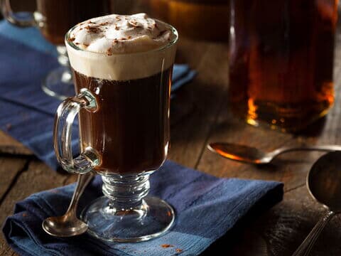 Ein Glas Irish Coffee mit cremiger Sahnehaube, serviert auf einem blauen Stofftuch, daneben ein Löffel auf einem rustikalen Holztisch
